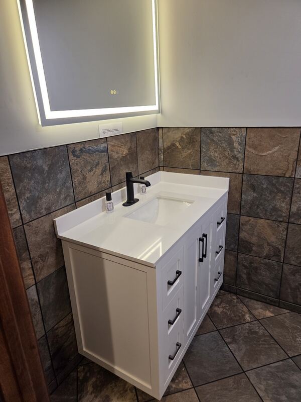 Finished bathroom vanity with white top, sink, black faucet, lighted mirror and stone-look tile.