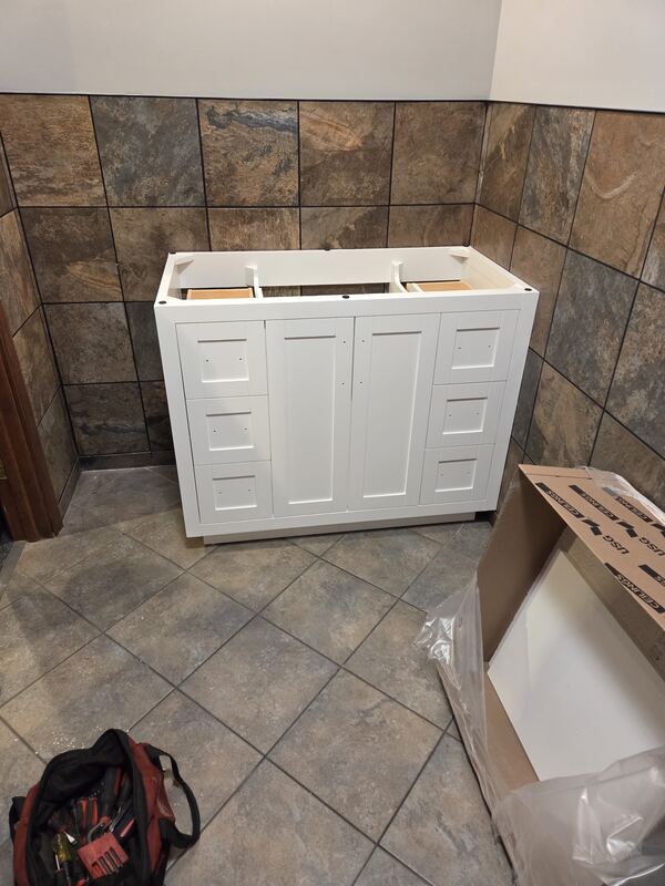 White vanity cabinet set in place against a stone-look tile wall. No top or hardware yet.